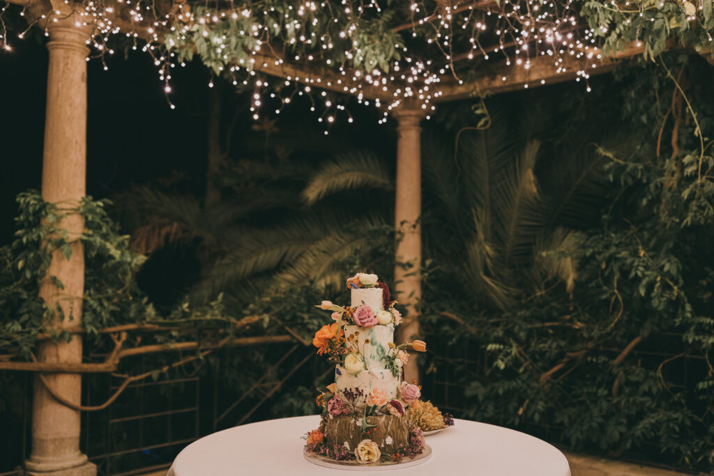 Tarta nupcial de cuatro plantas decorada con muchas flores y situada en una pérgola iluminada por un cielo estrellado. Fotografía de boda por Love Wanderers. Wedding Planner Martin & Gutfreund