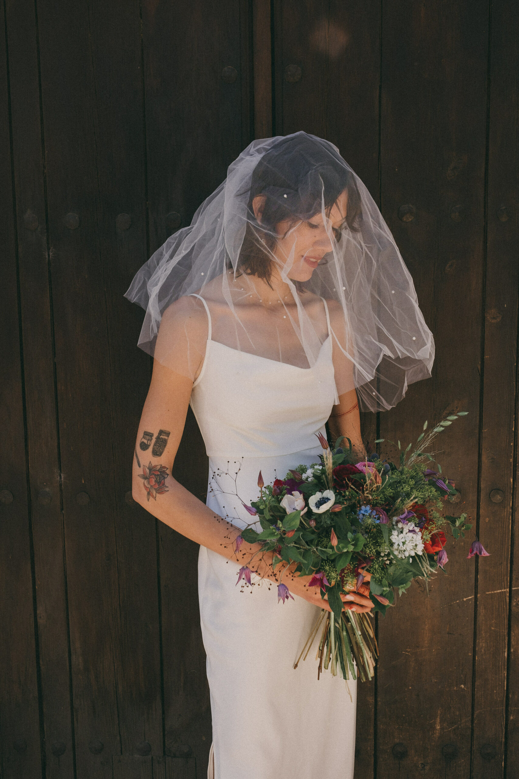 Una novia sujetando un ramo de flores frente a un portón de madera. Lleva un velo. Fotografía de boda por Love Wanderers. Wedding Planner Martin & Gutfreund